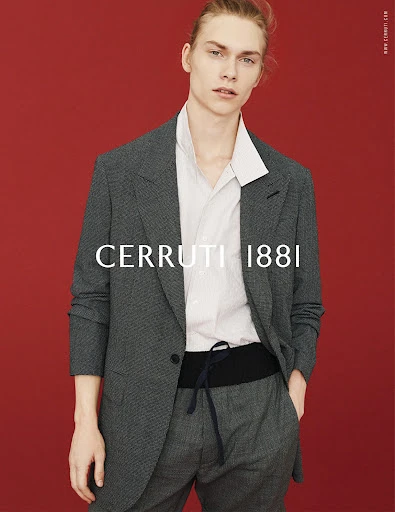 Fashion shot of a fair-skinned male model wearing a gray suit, white shirt, and black cummerbund against a red backdrop.