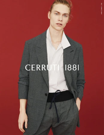 Fashion shot of a fair-skinned male model wearing a gray suit, white shirt, and black cummerbund against a red backdrop.