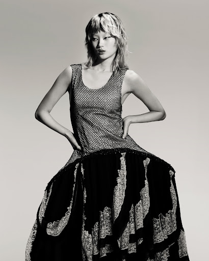 An Asian female model poses in a sleeveless silver top and layered black-and-white lace dress against a neutral backdrop.
