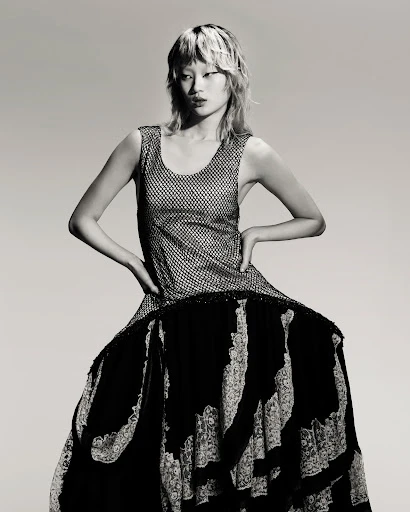 An Asian female model poses in a sleeveless silver top and layered black-and-white lace dress against a neutral backdrop.