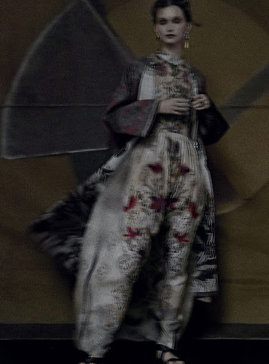 Woman in a patterned top and trousers, paired with a coat, poses in front of a blurred tan and gray backdrop in a fashion editorial.