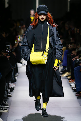 Runway fashion show featuring a model with red hair wearing a black coat, black baseball cap, and bright yellow bag and socks.