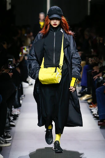 Runway fashion show featuring a model with red hair wearing a black coat, black baseball cap, and bright yellow bag and socks.