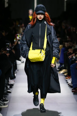 Runway fashion show featuring a model with red hair wearing a black coat, black baseball cap, and bright yellow bag and socks.