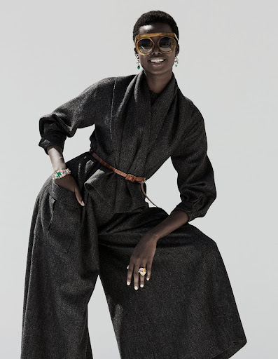 Fashionable African-American woman in gray wool suit and sunglasses poses against a bright background with hands on hips, smiling.