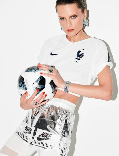 Female model in soccer jersey poses with a soccer ball in a studio setting, accessorized with statement jewelry and athletic wear.