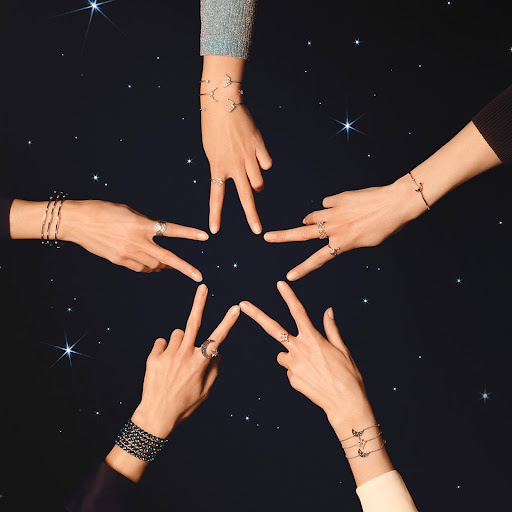 Five arms and hands converge toward the center of a dark star-speckled background, wearing rings and bracelets to form a star shape.