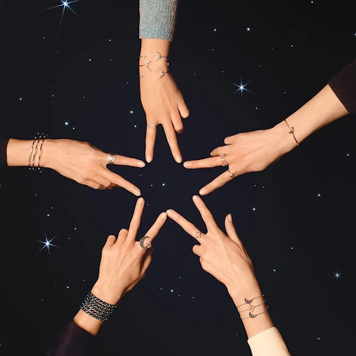 Five arms and hands converge toward the center of a dark star-speckled background, wearing rings and bracelets to form a star shape.