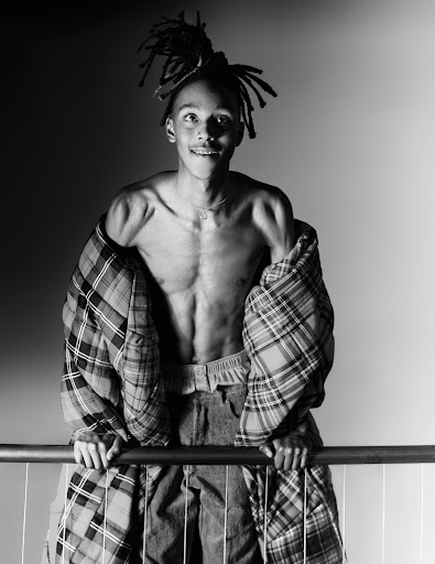 Monochrome portrait of a young man with dreadlocks draped in a plaid puffer jacket leaning on a railing.