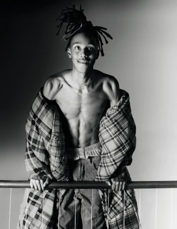 Monochrome portrait of a young man with dreadlocks draped in a plaid puffer jacket leaning on a railing.