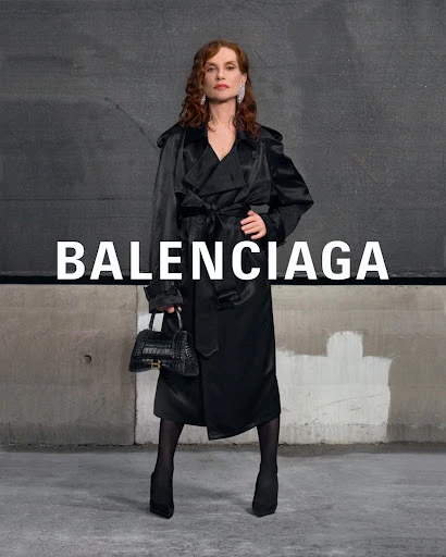 Adult female posing in a black trench coat, holding a black crocodile handbag, against a gray concrete wall with Balenciaga logo.