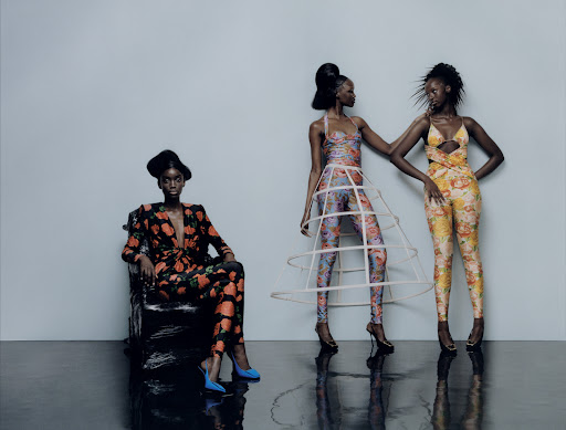 Three dark-skinned women in floral outfits pose in a studio setting. One woman sits on a chair, while the other two stand and interact.