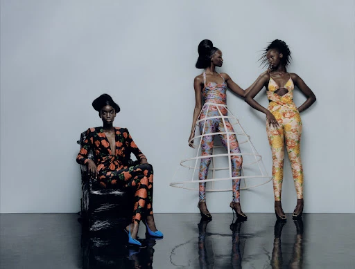 Three dark-skinned women in floral outfits pose in a studio setting. One woman sits on a chair, while the other two stand and interact.