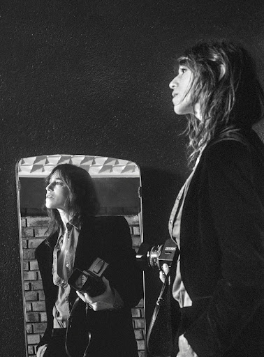 Monochrome portrait of a woman holding a vintage camera reflected in a mirror against a dark textured wall