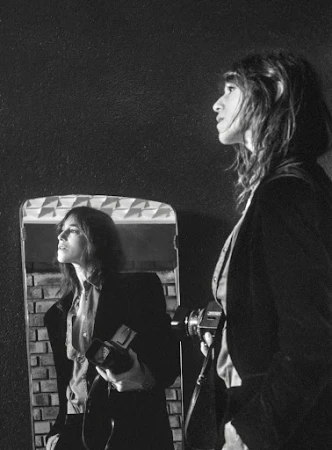 Monochrome portrait of a woman holding a vintage camera reflected in a mirror against a dark textured wall
