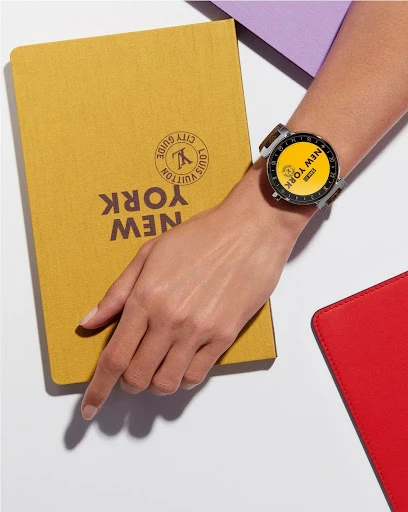Close-up studio shot featuring an adult's arm with a smartwatch next to Louis Vuitton City Guide book, with white background.