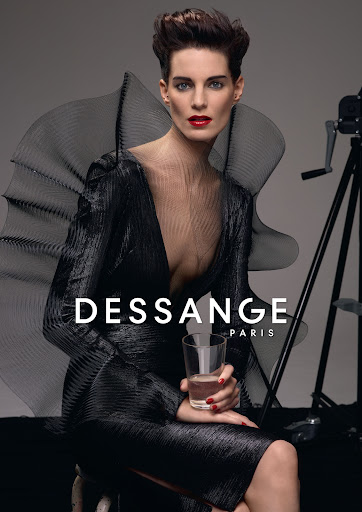 Elegant adult woman in a black dress with red lipstick holds a glass in a grey photo studio with a vintage camera in the background.