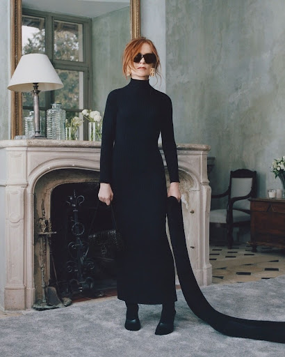 Woman in black turtleneck dress and sunglasses standing in an elegant interior with a fireplace and large mirror.