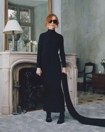 Woman in black turtleneck dress and sunglasses standing in an elegant interior with a fireplace and large mirror.