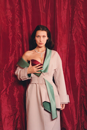 Bella Hadid poses in a studio setting wearing a robe and necklace. Red curtains hang in the background with a shallow depth of field.