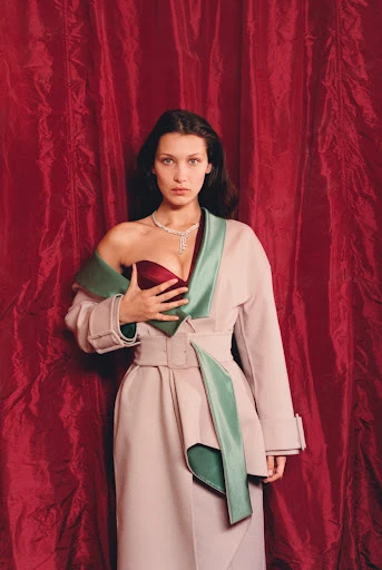 Bella Hadid poses in a studio setting wearing a robe and necklace. Red curtains hang in the background with a shallow depth of field.