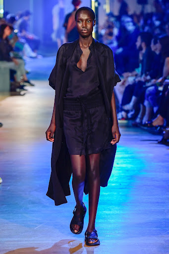 Runway fashion show featuring a Black female model in a black dress and sandals, walking in an indoor space with a crowd watching.