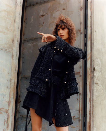 A stylish brunette adult woman in a fashion jacket and skirt poses in an industrial-style location, pointing into the distance.
