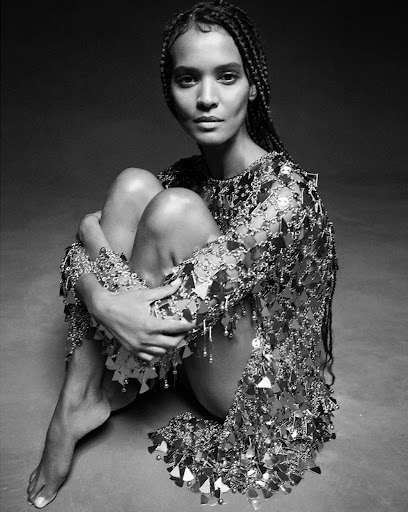 A Black woman with braided hair sits hugging her legs, posing in a shimmering sequin dress, set against a monochrome gray backdrop.