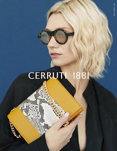 Fashion shot of a woman with blonde hair wearing sunglasses and a black top, holding a yellow purse with a snakeskin center panel.