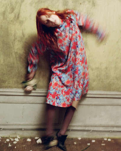 Redhead woman in floral dress seated with flowers, motion blur effect, against peeling wall, dark boots. Moody style, slightly desaturated.