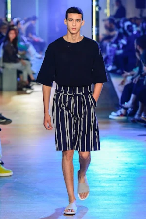 A male fashion model walks a runway wearing a dark top and striped shorts, presenting clothing in a fashion show, indoors with an audience.