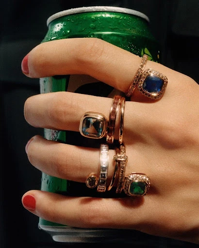 Close-up of a hand adorned with multiple rings, holding a green can with condensation, set against a dark background.