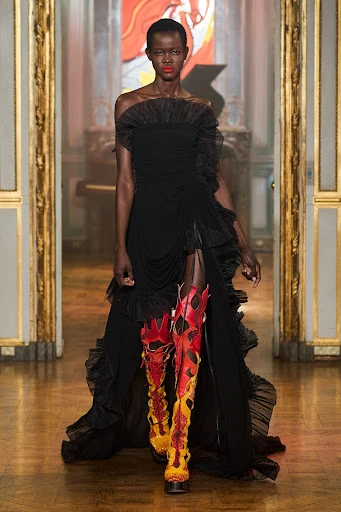 Runway fashion show featuring an African American fashion model in a black gown and flame-pattern boots, walking on the catwalk.