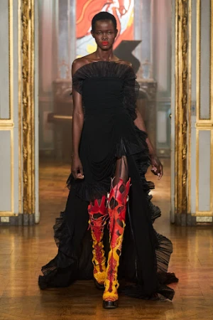 Runway fashion show featuring an African American fashion model in a black gown and flame-pattern boots, walking on the catwalk.