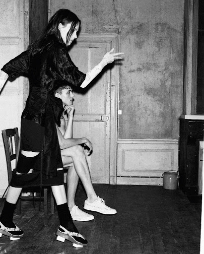 Monochrome fashion shot of a woman in a dark dress gesturing with her arm and a seated man in shorts and a white shirt in a room.