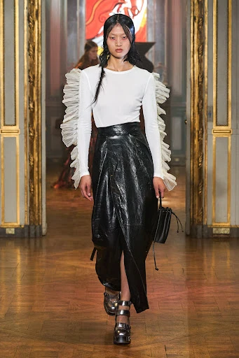 Runway fashion show featuring a model wearing a white top, black skirt, metallic platform shoes, and carrying a purse on a wooden floor.