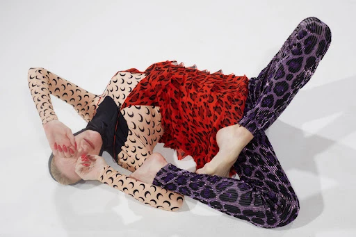 A model lies on a white background, wearing patterned clothing, including red and black top, crescent-print sleeves, and purple leopard-print pants.