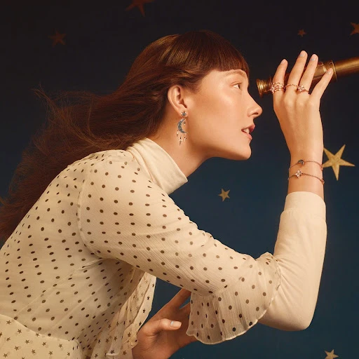 Elegant woman in a polka-dot blouse, looking through a telescope, adorned with jewelry against a navy star background for a fashion ad.