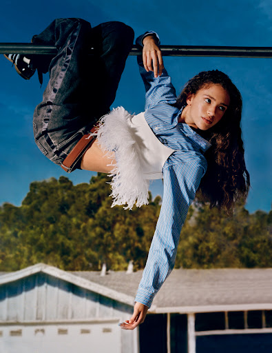 A person hangs upside down from a metal bar in a yard wearing jeans, an open striped shirt, and a white feathered top on a sunny day.