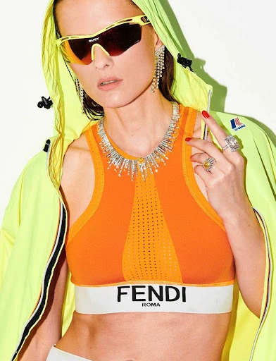 Stylish woman posing in an orange athletic crop top with a Fendi logo, statement jewelry, sunglasses, and neon green jacket.