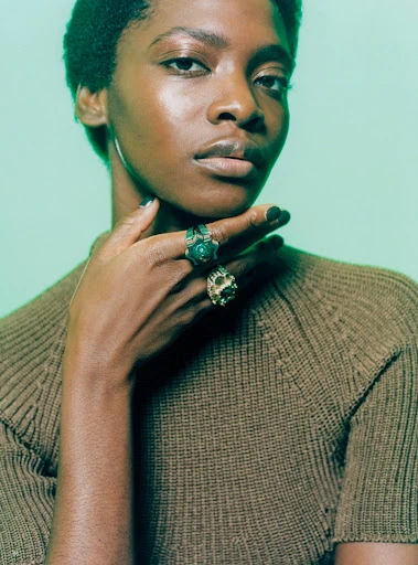 Portrait of an adult female wearing a brown knit sweater with green and gold rings on her fingers against a green gradient background.