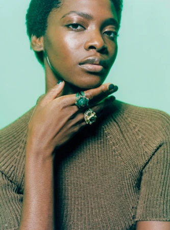 Portrait of an adult female wearing a brown knit sweater with green and gold rings on her fingers against a green gradient background.