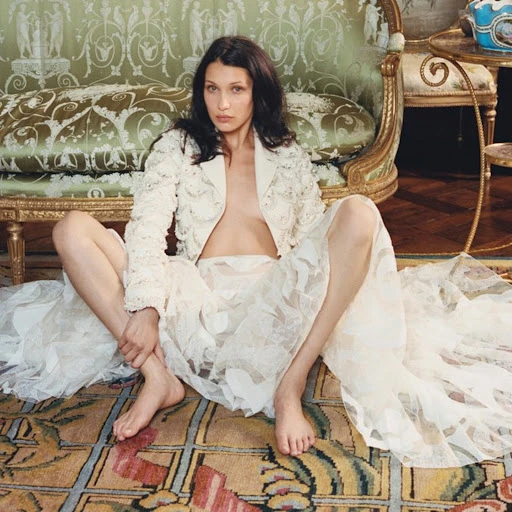 Brunette woman in a floral lace jacket and skirt sits barefoot on a patterned carpet in front of an ornate vintage couch.