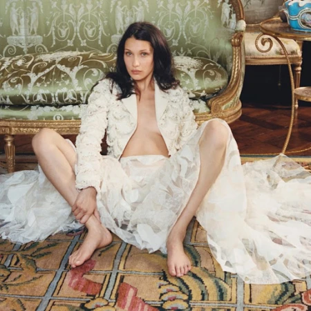 Brunette woman in a floral lace jacket and skirt sits barefoot on a patterned carpet in front of an ornate vintage couch.