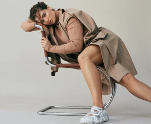 Model wearing a beige trench coat and ribbed top posing on a chrome cantilever chair against a plain white background.