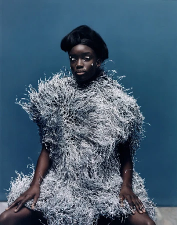 Black woman with dark voluminous hair wearing a textured silver fringe garment against a muted blue background