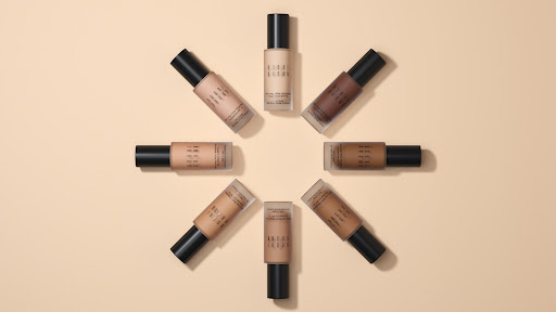 Multiple bottles of Bobbi Brown foundation arranged in a circular pattern on a light beige surface, showcasing various shades and tones.