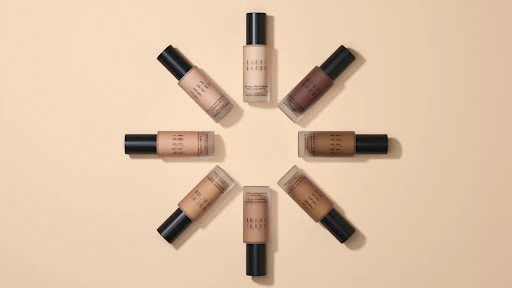 Multiple bottles of Bobbi Brown foundation arranged in a circular pattern on a light beige surface, showcasing various shades and tones.