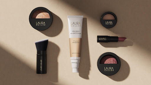 Overhead shot of Laura Geller makeup products including powder compacts, lipstick, makeup brush, and primer on a tan background with shadows.