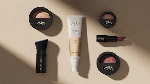Overhead shot of Laura Geller makeup products including powder compacts, lipstick, makeup brush, and primer on a tan background with shadows.